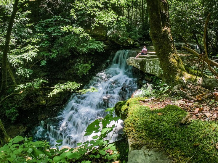 A woman sits on a rock ledge holding a camera above a large waterfall.