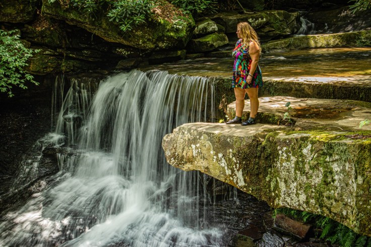 A woman wearing a colorful dress  stands on a rock ledge overlooking a waterfall