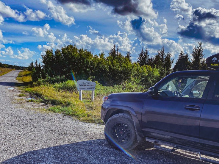 A navy 4runner in the foreground with a gray sign in the background that reads "Dolly Sods Wilderness." A dirt road stretches into the distance with blue skies and fluffy white clouds above, and green evergreen trees lining the road. 