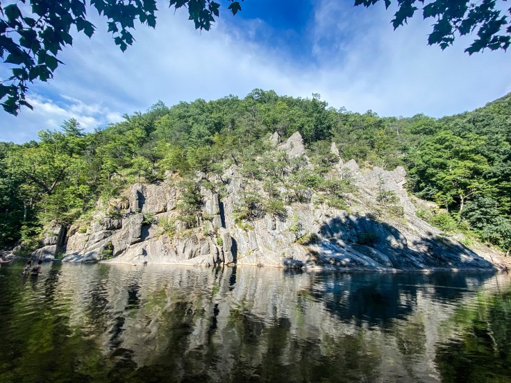 Dark water reflects in the foreground, with white/gray rock formations in the background framed by green confers and a blue sky with wispy clouds. 