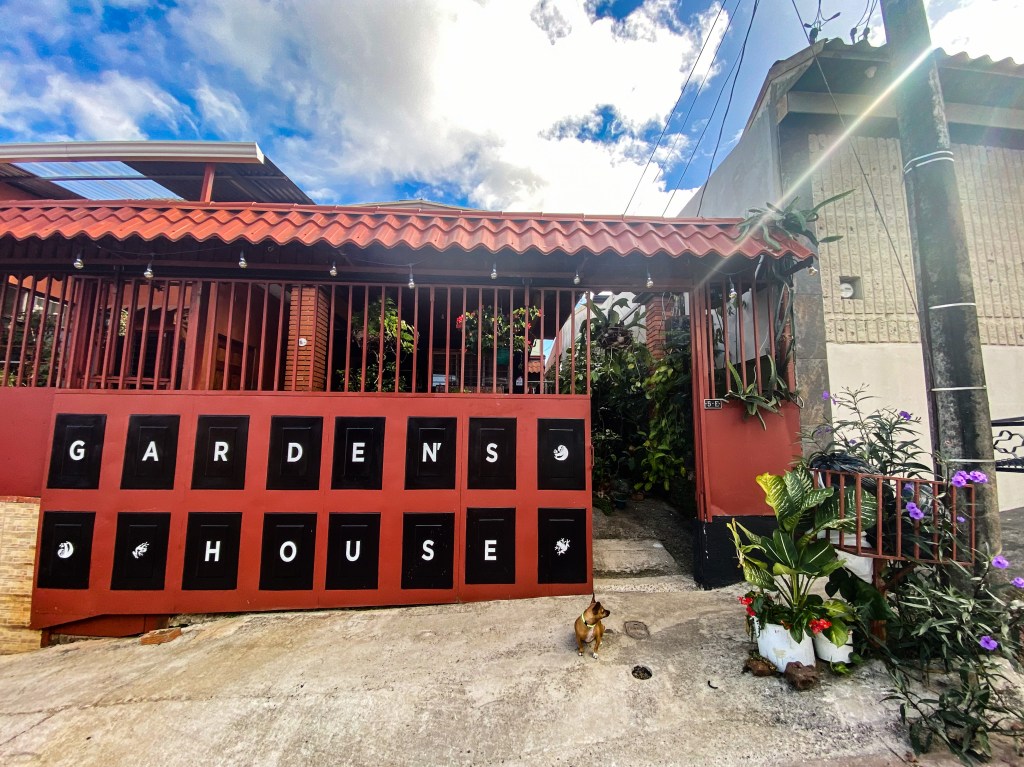 An orange gate with the words "GARDEN'S HOUSE" is in the background. Several green plants with purple and red flowers rame the right side of the image. A small brown dog sits in the foreground. 